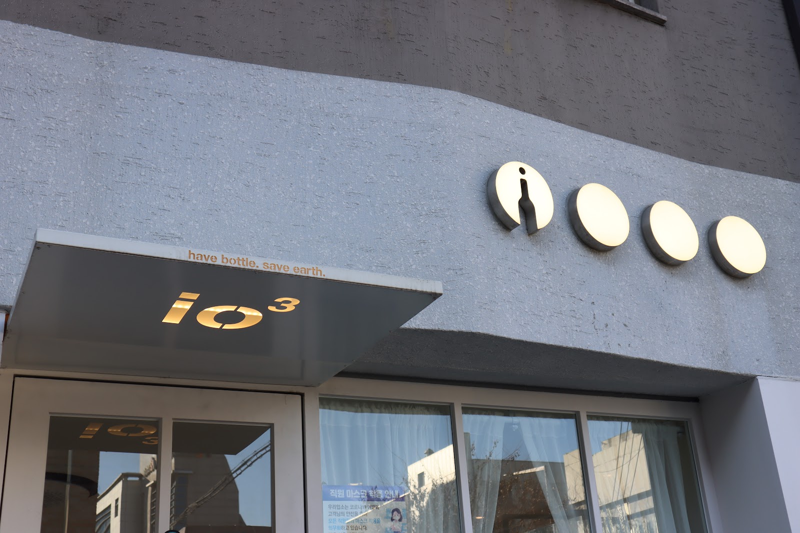 Cafe I/O³ — 外观:灰色抹灰面、圆形背光圆盘、"have bottle. save earth."宣言